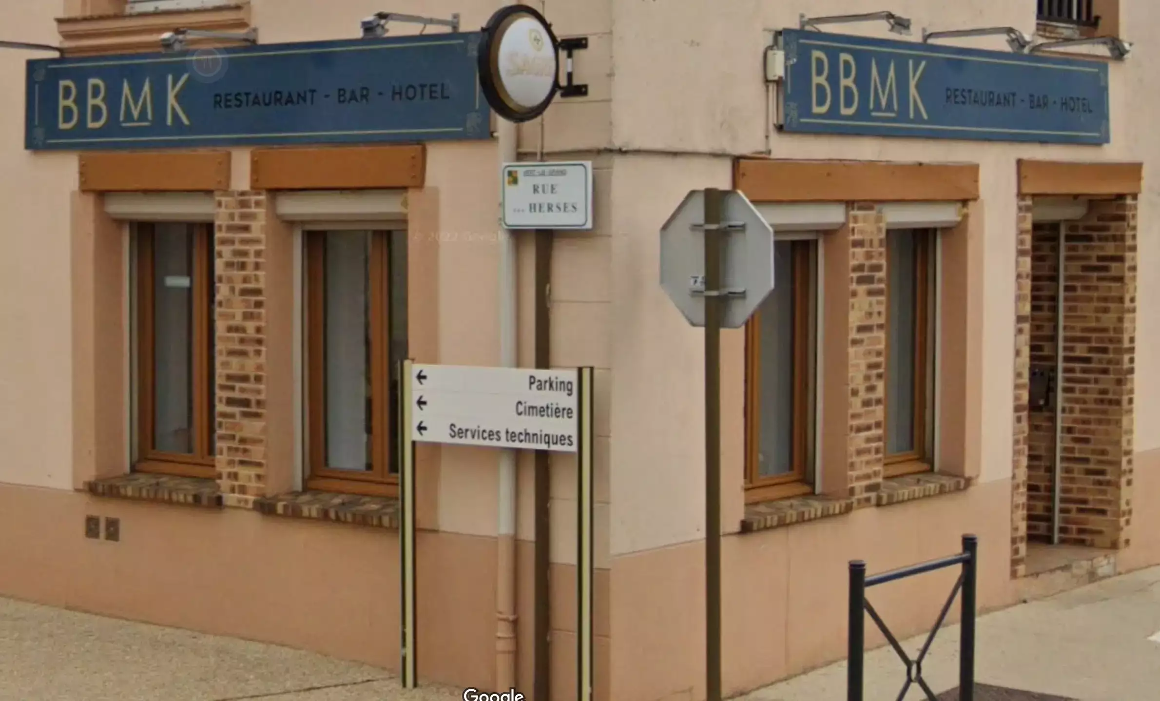 Odisourd-ou-dormir Façade d’un bâtiment portant l’enseigne ‘BBMK Restaurant - Bar - Hotel’, avec des fenêtres encadrées de briques et des volets en bois. Un panneau directionnel à proximité indique des lieux comme le parking, le cimetière et les services techniques. L’ensemble donne une impression de petit établissement local dans un environnement calme.